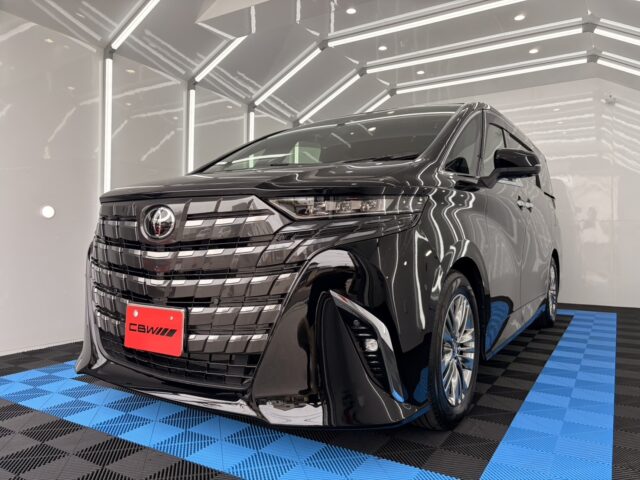 Shiny black minivan on a blue-and-black checkered showroom floor with bright geometric lighting overhead, viewed from the front-left.