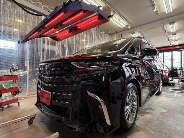 Black SUV in a car workshop under glowing red infrared curing lamps, with a red tool cart nearby.