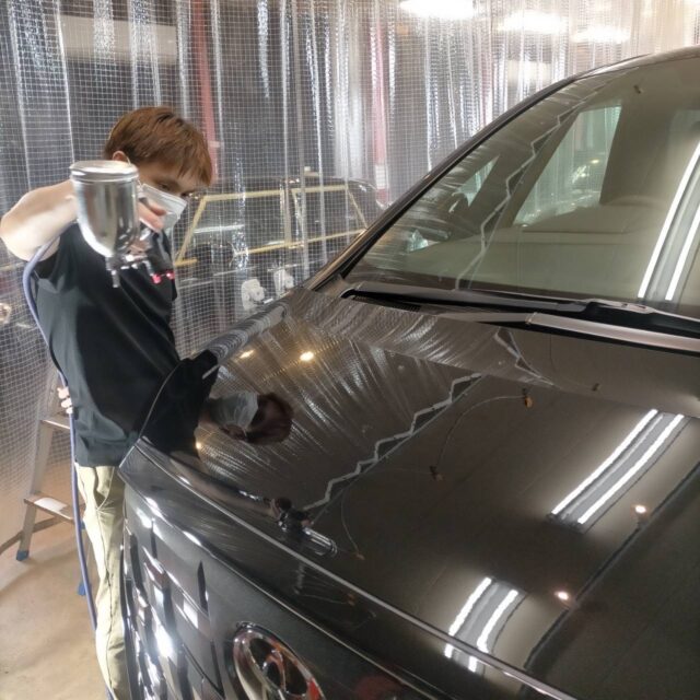 Worker wearing a mask and gloves sprays a dark car hood with a handheld spray gun in a workshop.