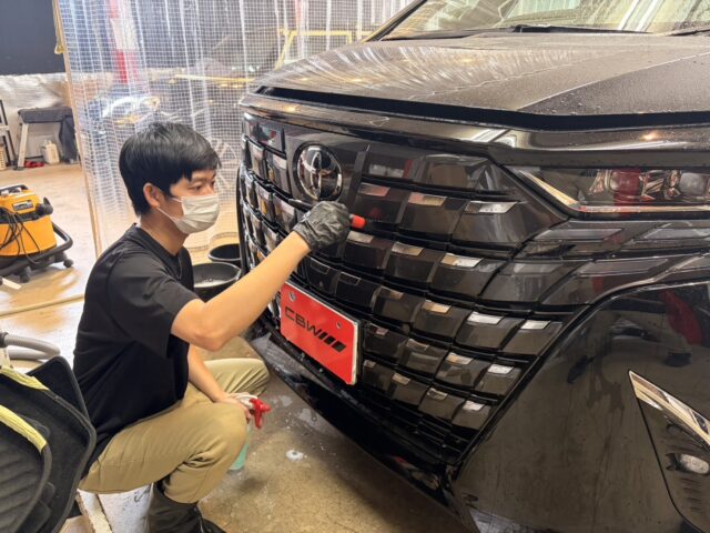 Detailer wearing a mask applies coating to the front grille of a black SUV in a workshop.