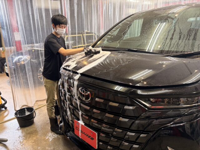 Man wearing a mask washes a black SUV, wiping the hood with soapy foam in a car wash bay.