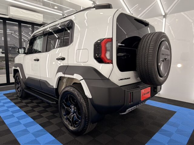 White off-road SUV (Land Cruiser) parked indoors, rear hatch open with spare tire mounted on the back, showing rugged gray bumper and black wheels.