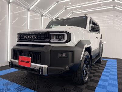 White Toyota pickup truck displayed in a bright showroom, front view showing a bold black grille, rugged bumper, and red license plate reading CBWII on a blue-and-black checkered floor.