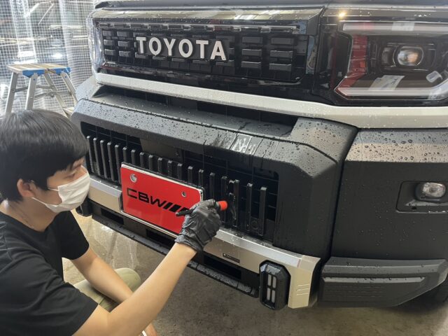 Man wearing a mask and gloves attaches a red license plate to the front bumper of a black Toyota truck in a workshop setting.