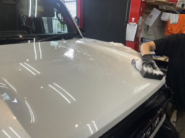 Person washing a white car hood with soap and a sponge in a car workshop/garage