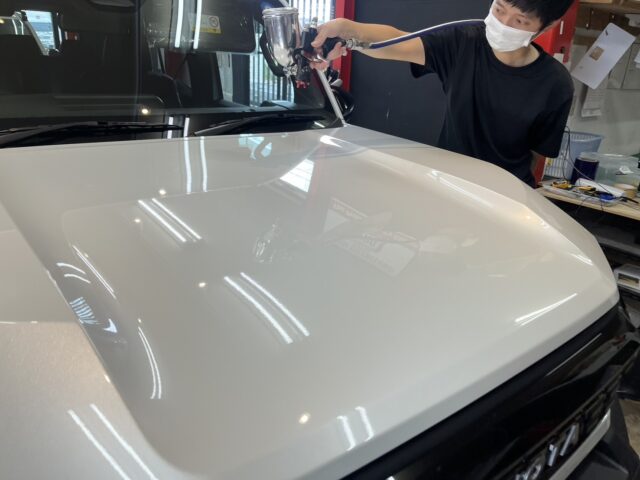 Person wearing a white mask polishes a white car hood with a spray gun in an auto body shop, reflected lights visible on the glossy surface.