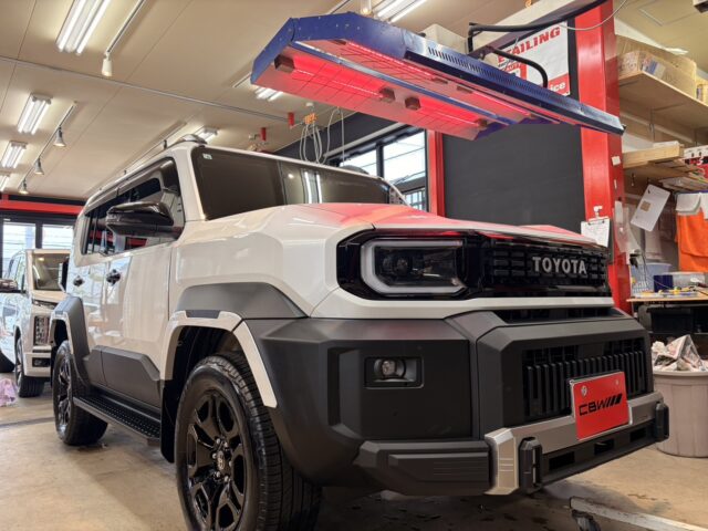 White Toyota SUV in a repair shop, front-left view under a blue and red vehicle hoist with tools and shelves in the background.