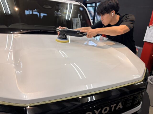 Person using a handheld polisher to buff the white Toyota SUV hood in a workshop.